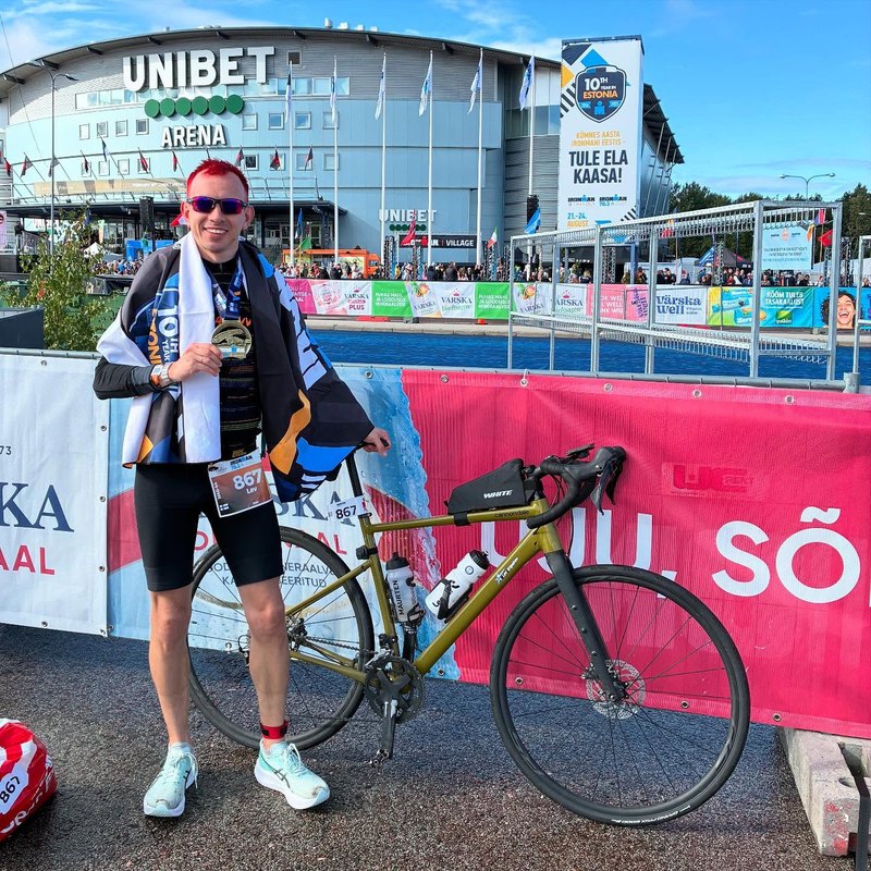 At the IRONMAN 70.3 Tallinn 2025 finish, wrapped in the Estonian flag, holding the medal next to the bike.